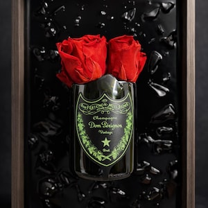 May include: A framed display featuring a Dom Pérignon champagne bottle with two vibrant red roses. The bottle is set against a black background with dark accents. The frame is a dark wood color.