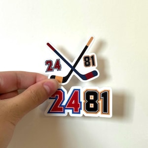 Op de afbeelding: Twee stickers met hockeythema. Eén sticker toont gekruiste hockeysticks met de nummers 24 en 81. De tweede sticker heeft de nummers 24 en 81 in een ander lettertype. De stickers zijn rood, blauw, zwart en bruin.
