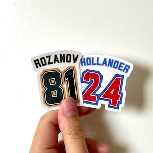 Könnte beinhalten: Zwei Aufkleber im Trikot-Stil. Einer zeigt den Namen "ROZANOV" über der Nummer "81" in Schwarz und Grün. Der andere zeigt "HOLLANDER" über der Nummer "24" in Rot und Blau. Die Aufkleber werden von einer Hand gehalten.