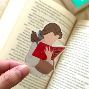 Peut inclure: Autocollant repr&eacute;sentant une femme de dessin anim&eacute; aux cheveux bruns lisant un livre rouge intitul&eacute; "A Court of Thorns and Roses". Elle porte une robe blanche et tient un panier marron. L'autocollant est pos&eacute; sur un livre ouvert.