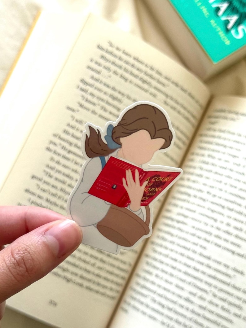 Peut inclure: Autocollant repr&eacute;sentant une femme de dessin anim&eacute; aux cheveux bruns lisant un livre rouge intitul&eacute; "A Court of Thorns and Roses". Elle porte une robe blanche et tient un panier marron. L'autocollant est pos&eacute; sur un livre ouvert.