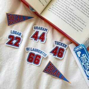 May include: A collection of stickers featuring sports jersey designs with names and numbers including Logan 22, Graham 44, Di Laurentis 66, and Tucker 46, alongside two triangular Briar U pennants and a blue Hockey Book Club ticket sticker.