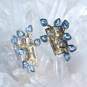 Puede incluir: Un par de pendientes de plata con un diseño de árbol dorado, cada uno adornado con piedras preciosas de color azul claro en forma de lágrima. Los pendientes se presentan sobre un fondo de cristales de cuarzo transparente.