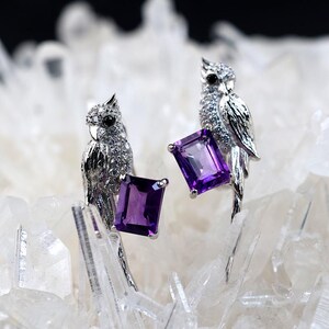 Puede incluir: Un par de pendientes de loro plateados. Cada pendiente presenta un diseño detallado de loro con acentos brillantes y una gran gema rectangular de color púrpura oscuro. Los pendientes están colocados sobre un fondo de cristales blancos.