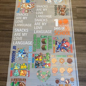 May include: A gray fabric banner with colorful illustrations and text. The banner features cartoon characters, cookies, and phrases like "Snacks are my love language" and "100 days of school".