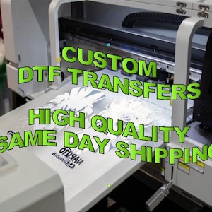 May include: A large white printer with the text "CUSTOM DTF TRANSFERS HIGH QUALITY SAME DAY SHIPPING" in green. The printer is printing a white design onto a white surface. The image is well-lit and shows the details of the printing process.
