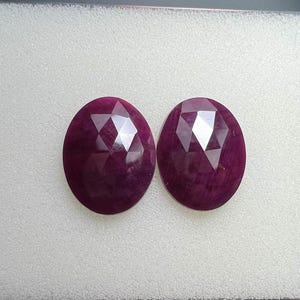 May include: Two oval-shaped, faceted gemstones with a deep red-purple hue. The gems are displayed on a white, textured surface within a black-edged tray. The facets create a sparkling effect, highlighting the stones' clarity and color.