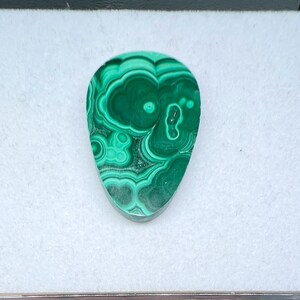 May include: A teardrop-shaped malachite gemstone with swirling patterns of dark and light green. The polished stone is set against a white background, showcasing its natural beauty and unique veining.
