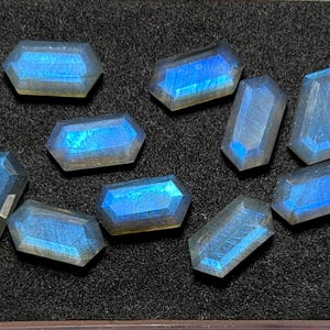 Pode incluir: Uma coleção de nove pedras preciosas de labradorite facetadas, cada uma com uma forma retangular distinta com bordas chanfradas. As pedras exibem um brilho azul iridescente vibrante em suas superfícies, contrastando com o fundo escuro.