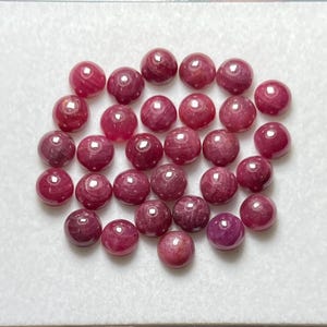 Pode incluir: Uma coleção de vinte e seis pequenas pedras preciosas redondas, lapidadas em cabochão, num tom magenta-rosa profundo. Cada pedra apresenta uma superfície lisa e polida com um brilho subtil, disposta num grupo sobre uma superfície branca.