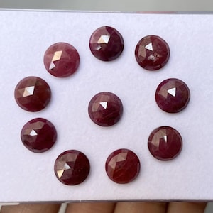 Könnte beinhalten: Eine Sammlung von zehn facettierten, runden Edelsteinen in verschiedenen tiefroten Farbtönen. Jeder Edelstein hat eine polierte, reflektierende Oberfläche mit mehreren Facetten. Die Edelsteine sind in einem kreisförmigen Muster auf weißem Hintergrund angeordnet.