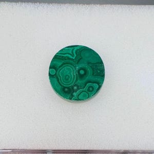 May include: A round, polished malachite stone with a vibrant green color and intricate swirling patterns. The stone is set against a white background, showcasing its natural beauty. This gemstone is ideal for jewelry making or collecting.