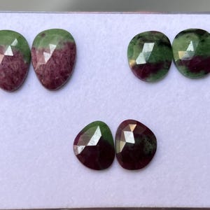 Op de afbeelding: Zes gefacetteerde edelstenen met een mix van groen en bordeaux. De edelstenen hebben verschillende vormen, waaronder druppel en ovaal. De edelstenen worden op een wit oppervlak gepresenteerd.