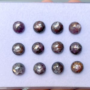 May include: Twelve faceted, round gemstones displayed in a clear plastic container. The gems are a dark, purplish-brown colour with reflective facets. The gems are arranged in three rows of four.