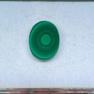 May include: Oval-shaped malachite gemstone with concentric circular patterns in varying shades of green. The polished stone is set against a white background, showcasing its natural beauty. This gemstone is ideal for jewelry making.