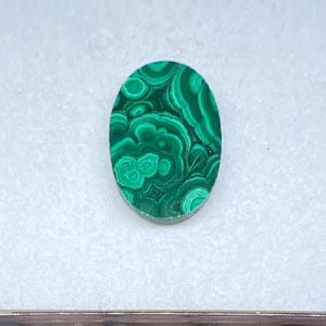 May include: Oval-shaped malachite gemstone with a vibrant green color and intricate swirling patterns. The polished stone is set against a white background, showcasing its natural beauty. This gemstone is ideal for jewelry making or collecting.