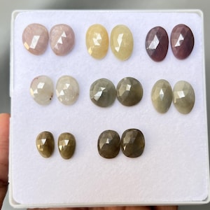 Könnte beinhalten: Eine Sammlung facettierter Edelsteine in verschiedenen Farben, darunter Rosa, Gelb, Grau und Braun. Die Edelsteine sind oval und birnenförmig und werden in einem weißen Tablett präsentiert. Die Edelsteine haben eine polierte, reflektierende Oberfläche.
