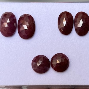 Peut inclure: Six pierres précieuses rouge foncé, taillées en ovale et rond, sont présentées sur une surface blanche. Les gemmes ont une surface polie et réfléchissante, captant la lumière. Les pierres sont disposées en deux rangées de trois, mettant en valeur leur couleur et leur taille.