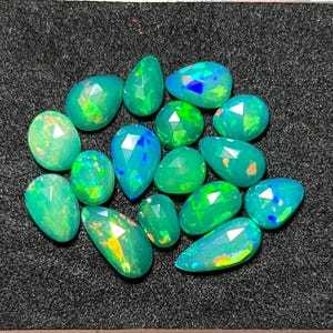 Puede incluir: Una colección de quince ópalos facetados, con forma de pera y ovalada, que muestran destellos vibrantes de color verde, azul y naranja. Cada piedra preciosa presenta una superficie pulida y multifacetada que refleja la luz.
