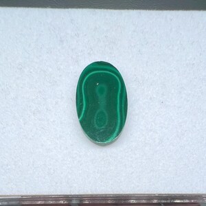 May include: Oval-shaped malachite gemstone with a vibrant green color and unique swirling patterns. The polished stone is set against a white background, showcasing its natural beauty. This gemstone is ideal for jewelry making.