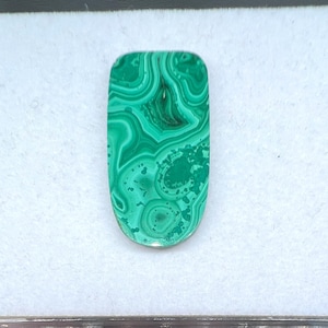 May include: A polished, oval-shaped malachite gemstone. The stone features a vibrant green color with swirling patterns and concentric circles, creating a unique and natural design. The gemstone is set against a white background.