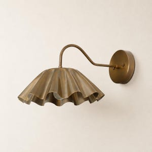 May include: A wall-mounted light fitting featuring a curved arm and a wide, scalloped metal shade in a brushed brass finish. The lamp includes a circular wall plate and a downward-facing shade with a wavy, pleated edge design in metallic gold.