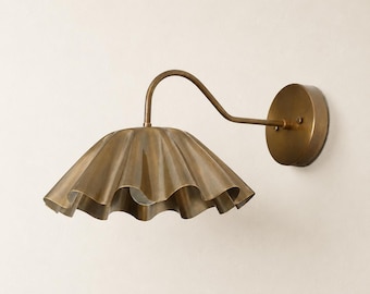 May include: A wall-mounted light fixture featuring a curved arm and a wide, scalloped metal shade in a brushed brass finish. The lamp includes a circular wall plate and a downward-facing shade with a wavy, pleated edge design in metallic gold.