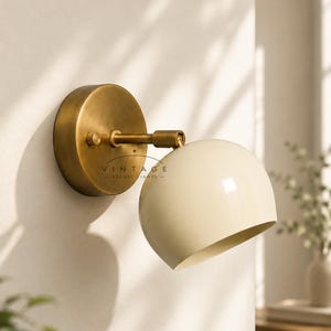 May include: A wall-mounted lamp featuring a circular brushed brass base and a smooth, cream-coloured dome-shaped shade. The fixture includes an adjustable arm and a small knob on the side. The text VINTAGE STARRY LIGHTS is visible on the base.