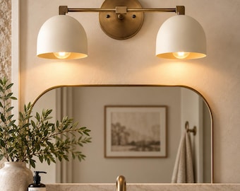 May include: A wall-mounted vanity light fixture featuring a horizontal brass bar and a circular backplate. Two matte off-white dome-shaped shades are attached to the ends, each housing a light bulb that emits a warm glow downward.