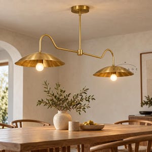 May include: A brass ceiling light fixture featuring two wide, shallow dome-shaped shades suspended from a central horizontal bar. Each shade houses a single glowing light bulb. The fixture has a metallic finish and a clean, linear structure.