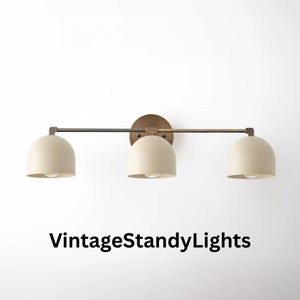 May include: A three-light wall fitting featuring a horizontal metal bar in a dark bronze finish with three matte cream-coloured dome shades. The fitting is mounted on a circular backplate. The text VintageStandyLights is visible below.