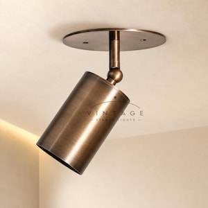 May include: A cylindrical bronze-toned spotlight fixture with a circular ceiling mount and an adjustable swivel joint. The metal surface features a brushed finish. The text VINTAGE STANOY LIGHTS is printed on the side of the cylindrical light housing.