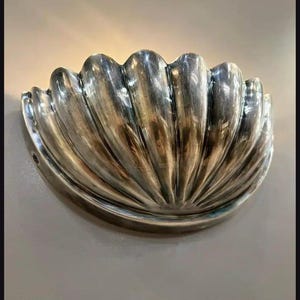 Puede incluir: Una lámpara de pared plateada con forma de concha. La lámpara tiene un diseño festoneado con crestas verticales, creando una superficie texturizada. El acabado metálico refleja la luz, resaltando las curvas y los detalles.