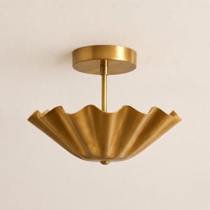 May include: A brass ceiling light fitting featuring a circular canopy, a central vertical rod, and a wide, bowl-shaped shade with a scalloped, wavy edge. The metal has a smooth, polished finish that reflects light across its curved, fluted surface.