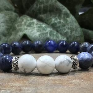 May include: A beaded bracelet featuring a combination of dark blue, white, and silver beads. The bracelet has a central section of three white beads, flanked by dark blue and silver accent beads.