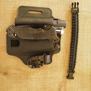 Peut inclure: Un étui multi-outils en cuir noir avec un couteau pliant, une lampe de poche et un porte-clés. Un bracelet de survie en paracorde noire est à côté de l'étui. Les articles sont sur une surface en bois brun clair.