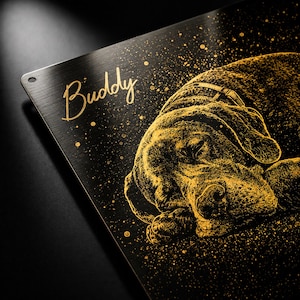 May include: A black metal sign featuring a gold illustration of a sleeping dog. The dog wears a collar. The name "Buddy" is written in gold script above the dog. Gold speckles are scattered across the sign.