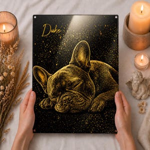 Puede incluir: Un letrero de metal negro con una ilustración dorada de un bulldog francés durmiendo. El nombre del perro, "Duke", está escrito en oro. El fondo es negro con motas doradas. El letrero está sostenido por manos.