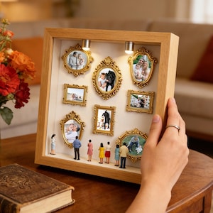 Könnte beinhalten: Eine Holzrahmen-Ausstellung mit neun verzierten goldfarbenen Bilderrahmen, die jeweils ein Foto enthalten. Miniaturfiguren stehen unter den Rahmen. Zwei kleine Lichter beleuchten die Ausstellung. Eine Hand hält den Rahmen auf einem Holztisch.