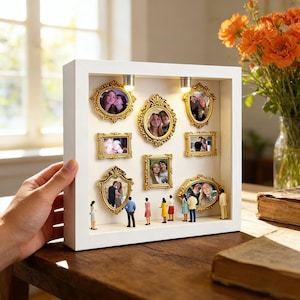 May include: A white shadow box with nine gold-framed photos and miniature figures. The box has two small lights at the top. The photos feature various people. The box is held by a person.