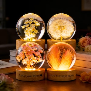 Puede incluir: Cuatro esferas de vidrio iluminadas con inclusiones florales y vegetales, cada una sobre una base de madera. Las esferas contienen margaritas amarillas, un diente de león, flores rosas y plumas naranjas. Dos bases dicen "Love you Mom" y "FOR THE BEST MOM".