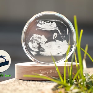 Peut inclure: Un souvenir en cristal clair et facetté, représentant une échographie d'un bébé. Le cristal est posé sur un socle en bois gravé du nom "Baby Ben". Une boîte cadeau avec un ruban bleu est également visible.