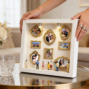 Puede incluir: Una caja de sombra blanca con múltiples fotos enmarcadas en oro de parejas y familias. Pequeñas figuras están de pie frente a las fotos. Dos pequeñas luces están en la parte superior. La caja es sostenida por una persona.