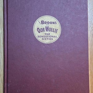 May include: A burgundy hardcover book with a circular emblem. The emblem reads "The Broons and Oor Wullie The Sensational Sixties" in gold lettering. The book is closed and lies on a wooden surface.