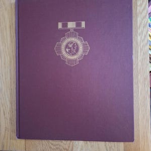May include: A maroon book featuring a gold emblem and text. The emblem is a circular design with a ribbon at the top. The text reads "For God, The Street and The Empire". The book rests on a wooden surface.
