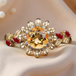 Op de afbeelding: Een goudkleurige ring met een grote, ronde, gele edelsteen in het midden, omringd door kleinere witte stenen en bloemblaadjes. De band heeft blad- en rode edelsteen details.