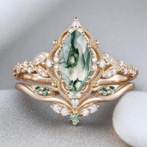 May include: A gold-toned engagement ring set featuring a large, oval-cut green gemstone with a unique pattern. The ring is adorned with small, clear accent stones and delicate leaf-like details. The set includes a matching band with green gemstones.