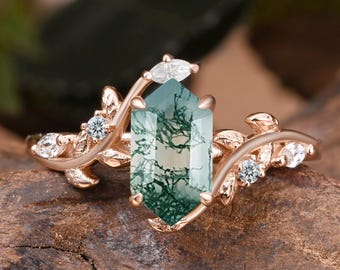 May include: Rose gold ring featuring a large, hexagonal, green moss agate gemstone. The ring has a nature-inspired design with leaf and small diamond accents. The ring is set on a wooden surface.