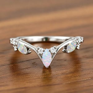 May include: A V-shaped silver ring featuring three marquise-cut opals and four small round clear stones. The central opal is positioned at the point of the V, with one opal on each side, accented by small clear stones set into the metal band.