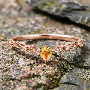 May include: A rose gold ring featuring a central pear-shaped orange gemstone flanked by two small round clear stones. The band is crafted with a detailed leaf-like vine pattern that extends along both sides of the central setting.